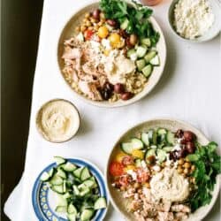 Two bowls of Mediterranean-style salad with couscous, shredded chicken, cherry tomatoes, cucumber, olives, parsley, feta, and hummus on a white tablecloth, surrounded by side dishes.