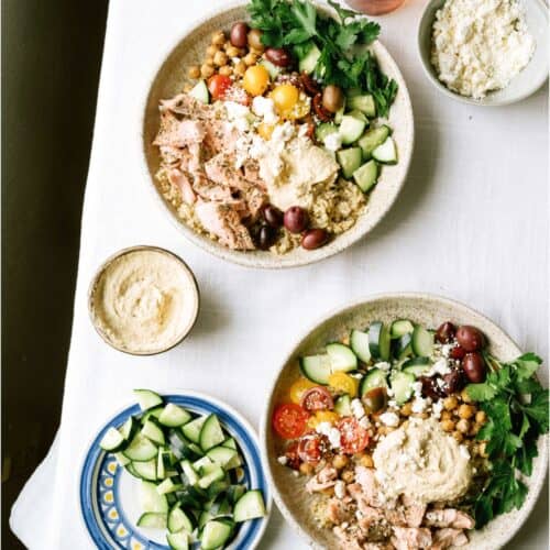 Two bowls of Mediterranean-style salad with couscous, shredded chicken, cherry tomatoes, cucumber, olives, parsley, feta, and hummus on a white tablecloth, surrounded by side dishes.