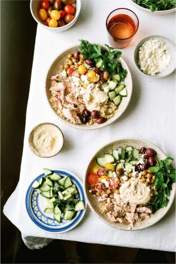 Two bowls of Mediterranean-style salad with couscous, shredded chicken, cherry tomatoes, cucumber, olives, parsley, feta, and hummus on a white tablecloth, surrounded by side dishes.