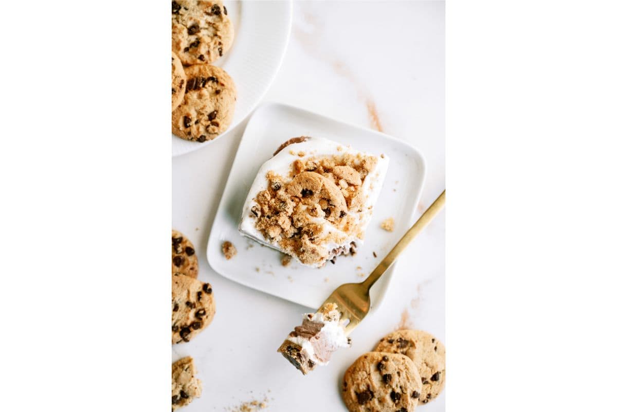 A square slice of dessert topped with crumbled chocolate chip cookies on a white plate, surrounded by whole cookies and a gold fork with a bite.