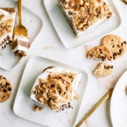Two dessert squares topped with crumbled cookies are served on white plates with gold forks. Whole and broken chocolate chip cookies are scattered around.