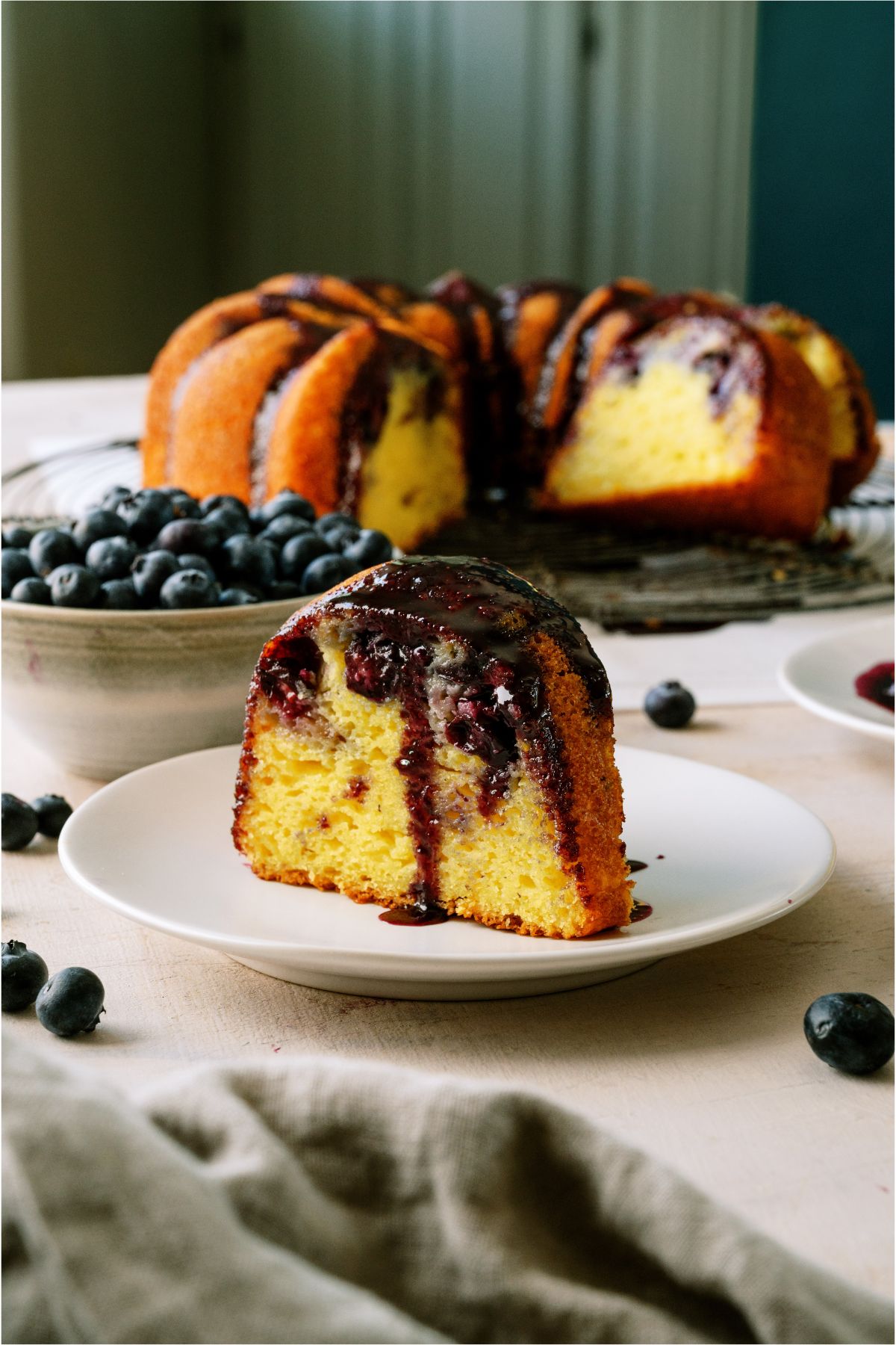 A slice of Lemon Blueberry Bundt Cake on a plate with the remaining cake in the background