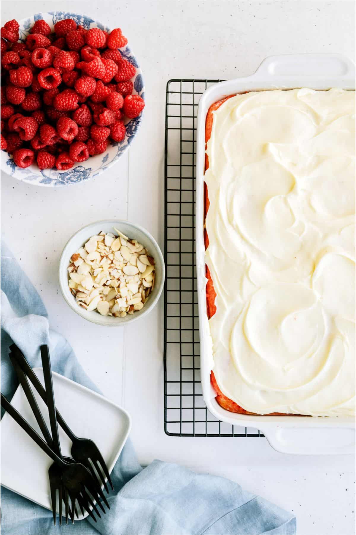 White Raspberry Poke Cake frosted with a small bowl of fresh berries and a small bowl of sliced almonds off to the side.