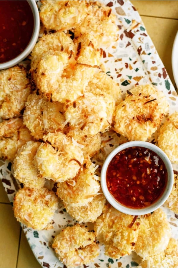 A platter of golden-brown coconut shrimp arranged around two small bowls of red dipping sauce.