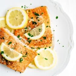 Two pieces of cooked salmon garnished with lemon slices and chopped herbs are served on a white plate.