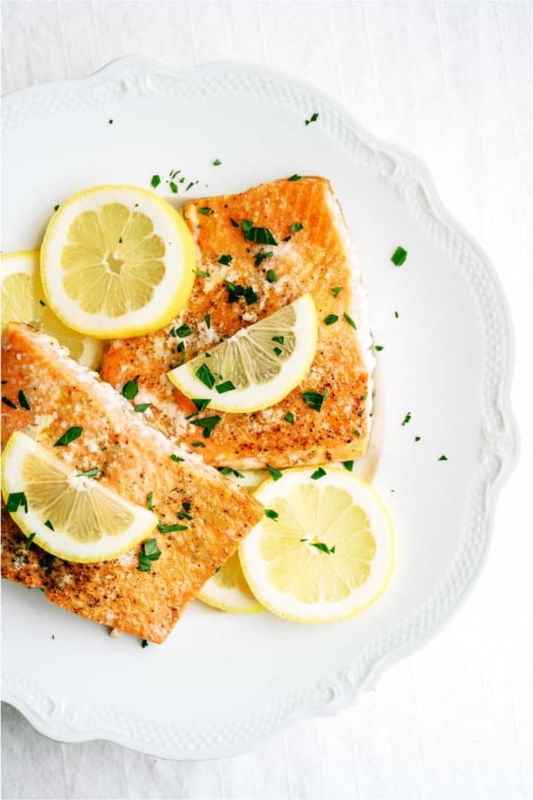 Two pieces of cooked salmon garnished with lemon slices and chopped herbs are served on a white plate.