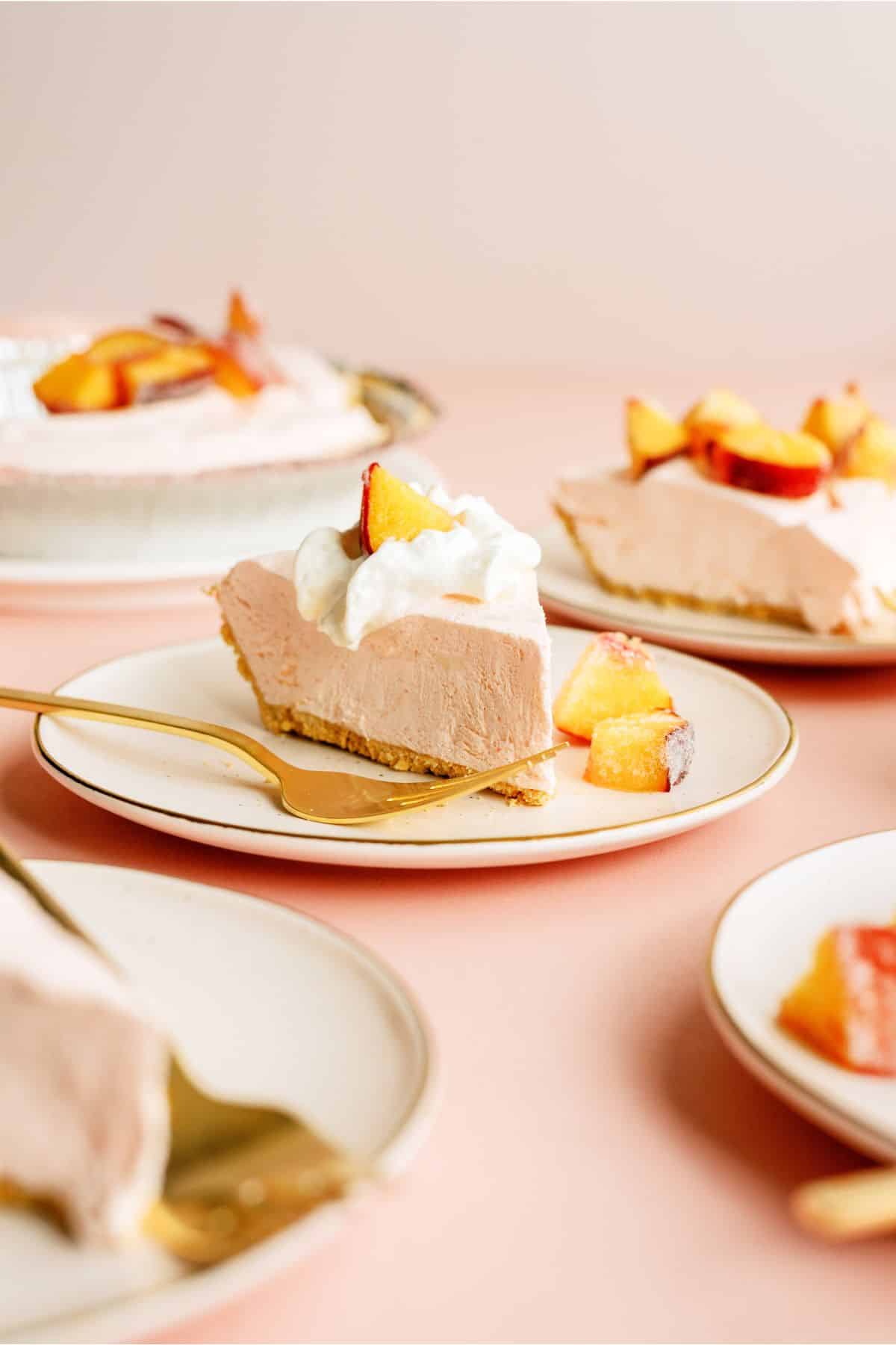 A slice of creamy pie topped with whipped cream and peach pieces on a plate with a gold fork; other slices are visible in the background.