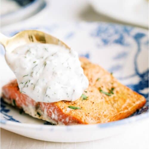 A spoonful of creamy dill sauce is being poured onto a cooked salmon fillet on a decorative plate.