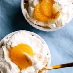 Two bowls of creamy dessert topped with mandarin orange slices, banana pieces, pineapple chunks, and mini marshmallows, served with gold-colored spoons on a blue surface.