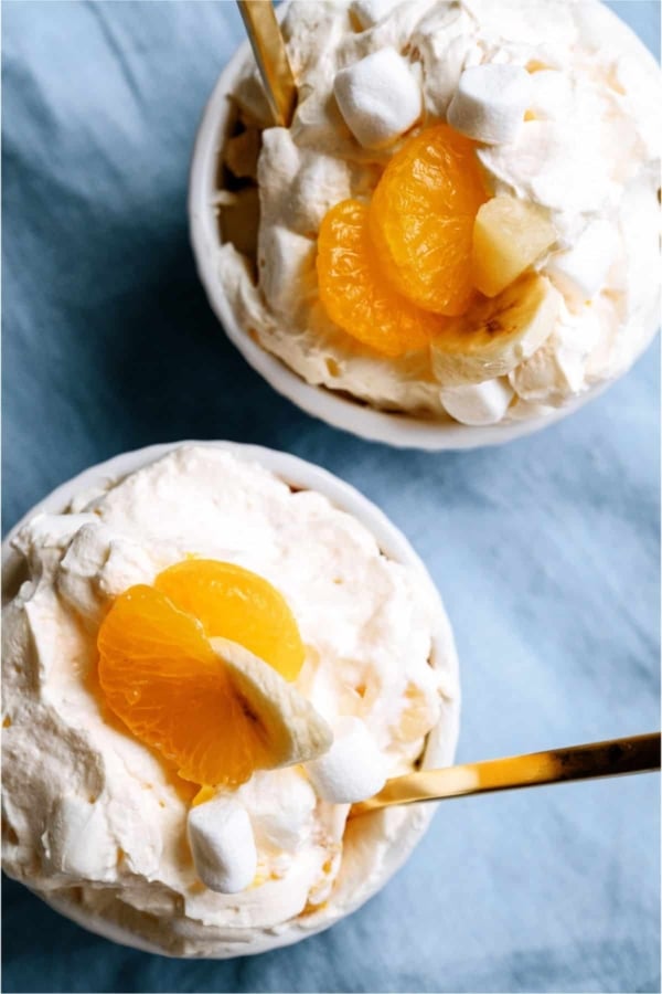 Two bowls of creamy dessert topped with mandarin orange slices, banana pieces, pineapple chunks, and mini marshmallows, served with gold-colored spoons on a blue surface.