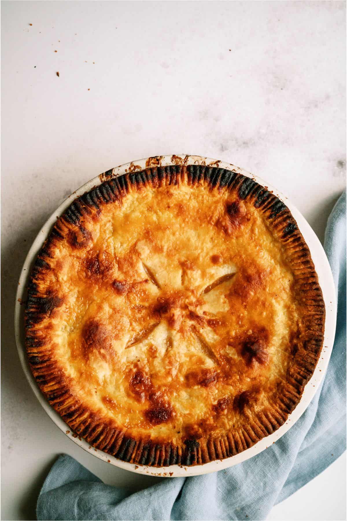 A golden-brown pie with a crimped edge and decorative vent in the center sits on a white surface next to a light blue cloth.
