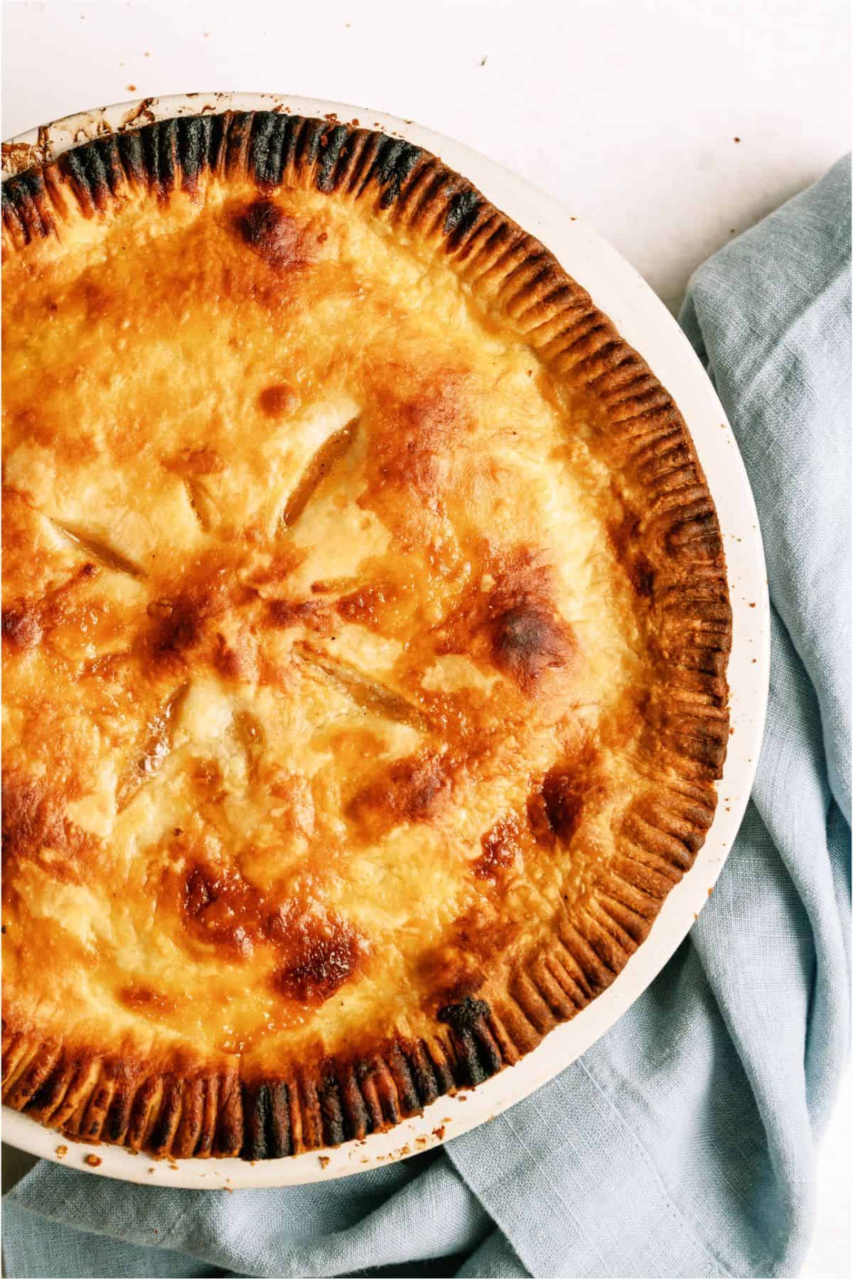 A golden-brown pie with a flaky crust sits in a white dish, next to a light blue cloth.
