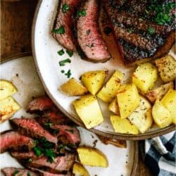 Two plates with sliced steak, roasted potato chunks, and a garnish of chopped herbs on a wooden table.