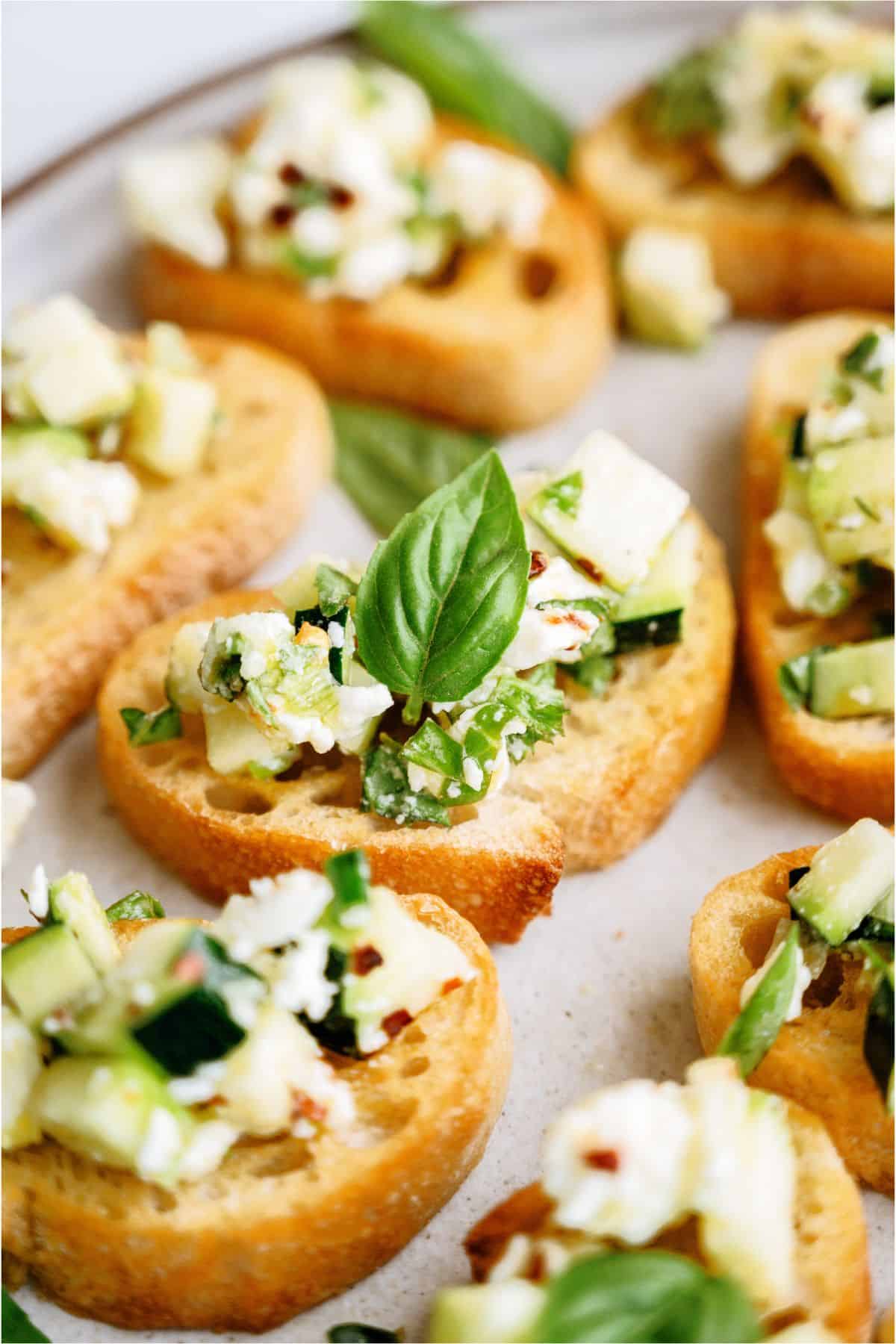 Crostini topped with diced cucumber, feta, herbs, and a basil leaf, arranged on a plate.