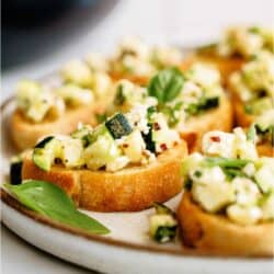 Slices of bread topped with a mixture of diced zucchini, melted cheese, and herbs, garnished with basil leaves on a ceramic plate.