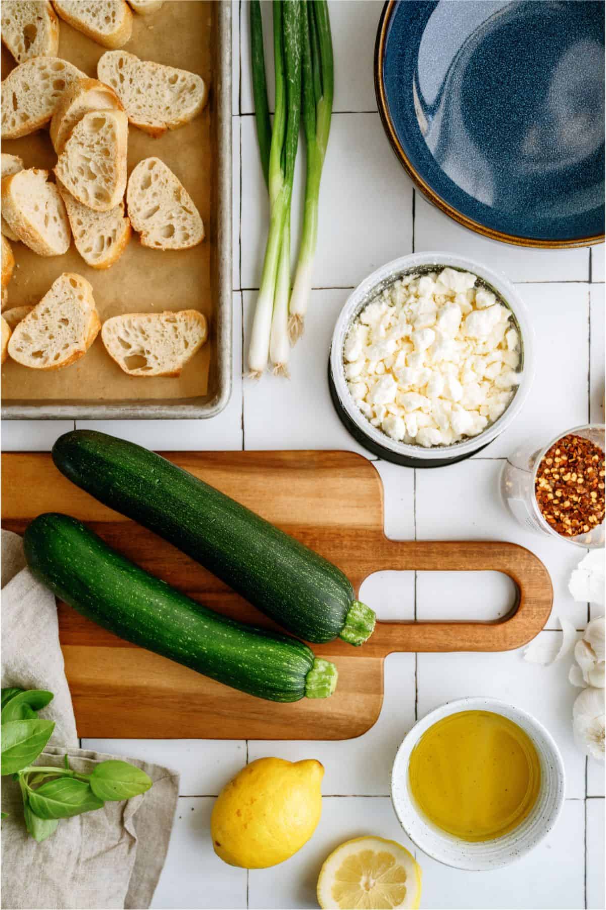 Top view of cooking ingredients: sliced bread, green onions, zucchini, feta cheese, red pepper flakes, basil, lemon, and a bowl of olive oil on a white surface.
