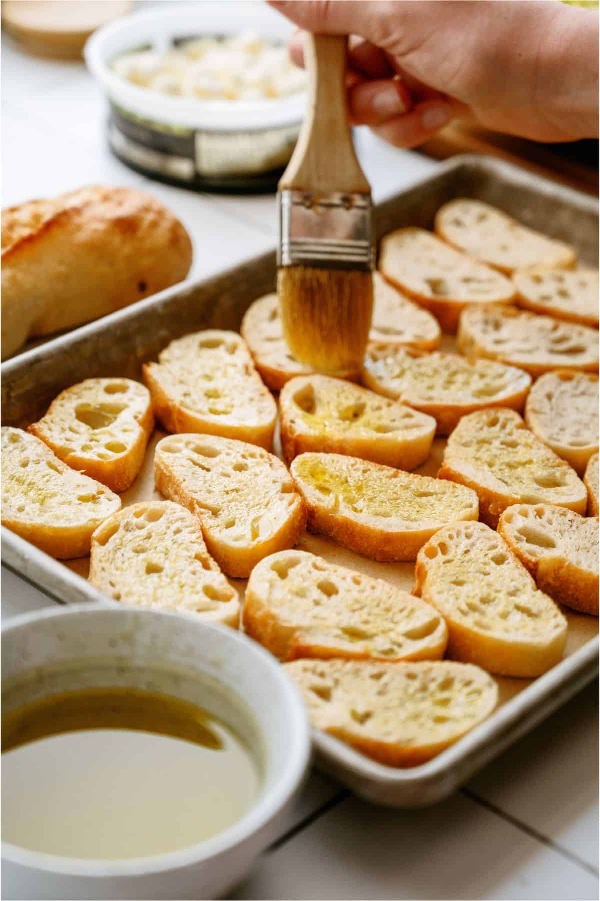 A person brushes olive oil on slices of bread arranged on a baking sheet, with a bowl of olive oil nearby.