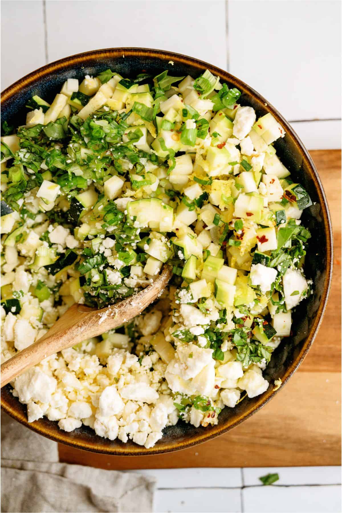 A bowl filled with diced zucchini, herbs, and crumbled feta cheese, with a wooden spoon resting inside.