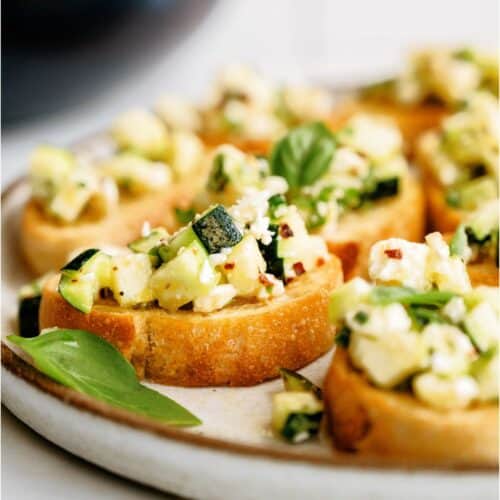 Slices of bread topped with a mixture of diced zucchini, melted cheese, and herbs, garnished with basil leaves on a ceramic plate.