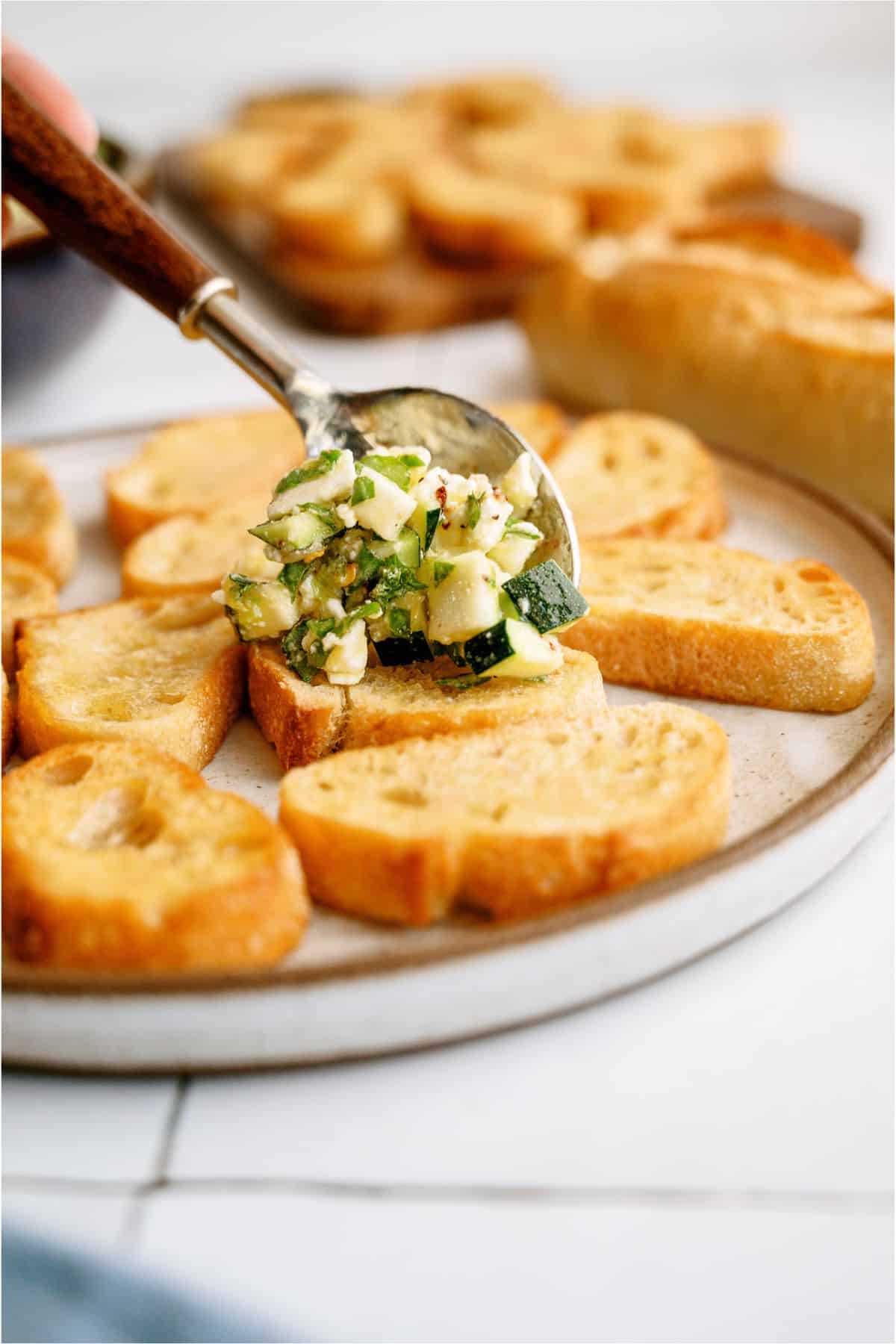 A spoonful of vegetable topping is being placed on toasted bread slices arranged on a plate.