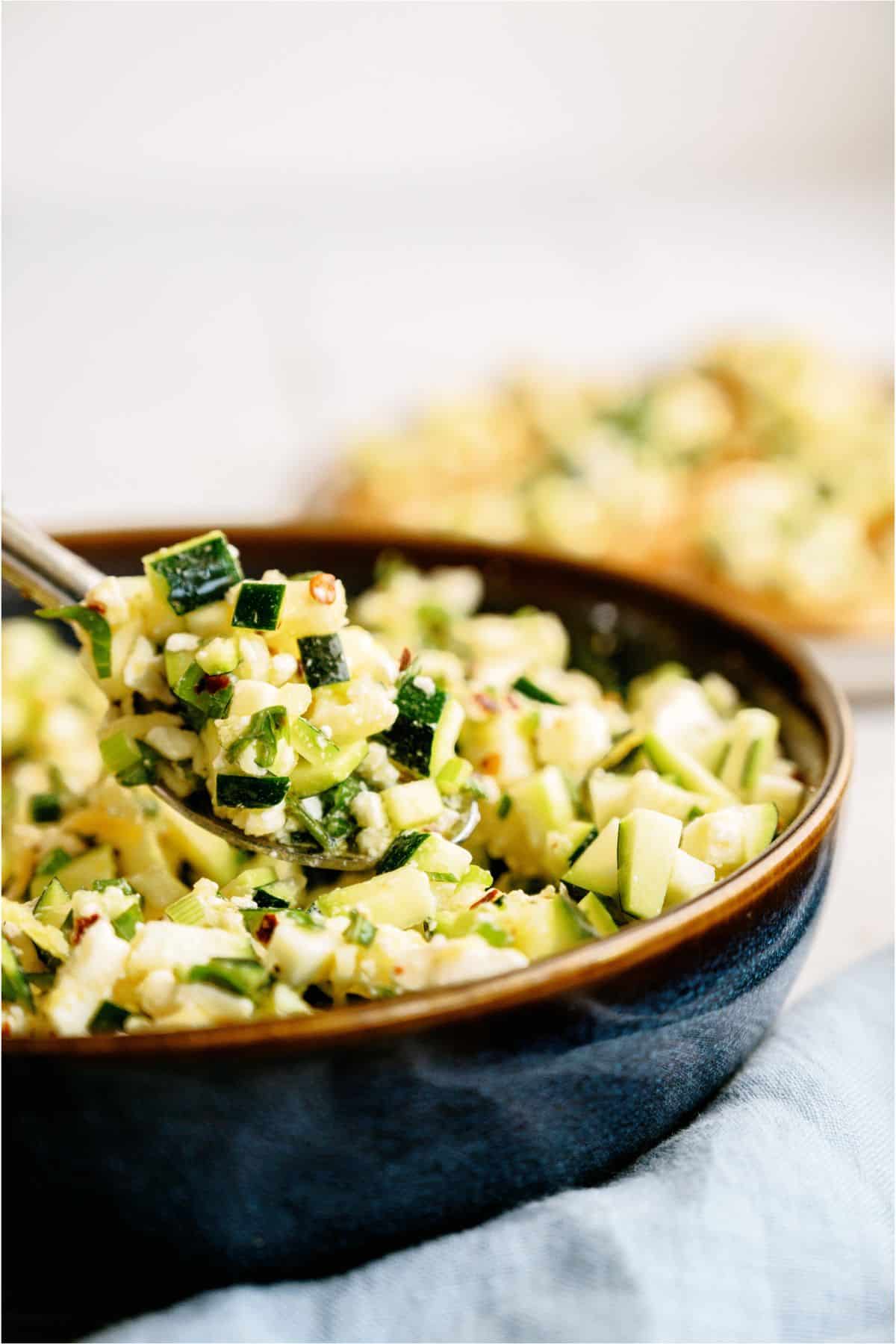 Close-up of a bowl filled with a salad of chopped zucchini and corn, with a spoon lifting a portion.