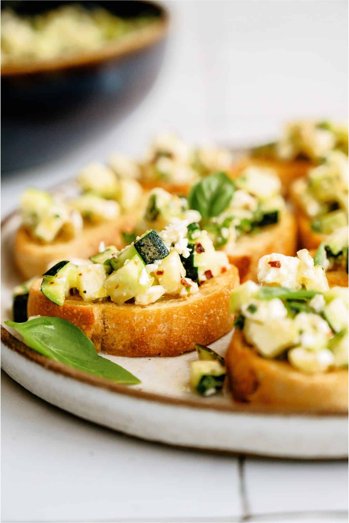 Slices of bread topped with a mixture of diced zucchini, melted cheese, and herbs, garnished with basil leaves on a ceramic plate.