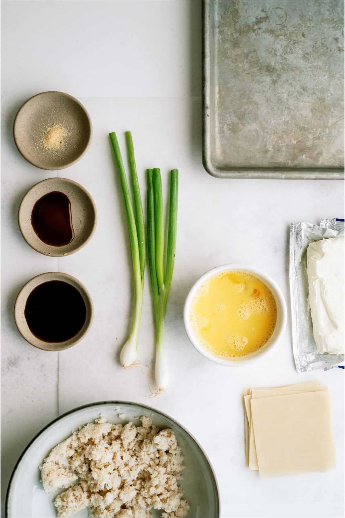 Ingredients on a countertop: ground meat, cream cheese, egg wash, soy sauce, garlic powder, green onions, wonton wrappers, and a baking tray.