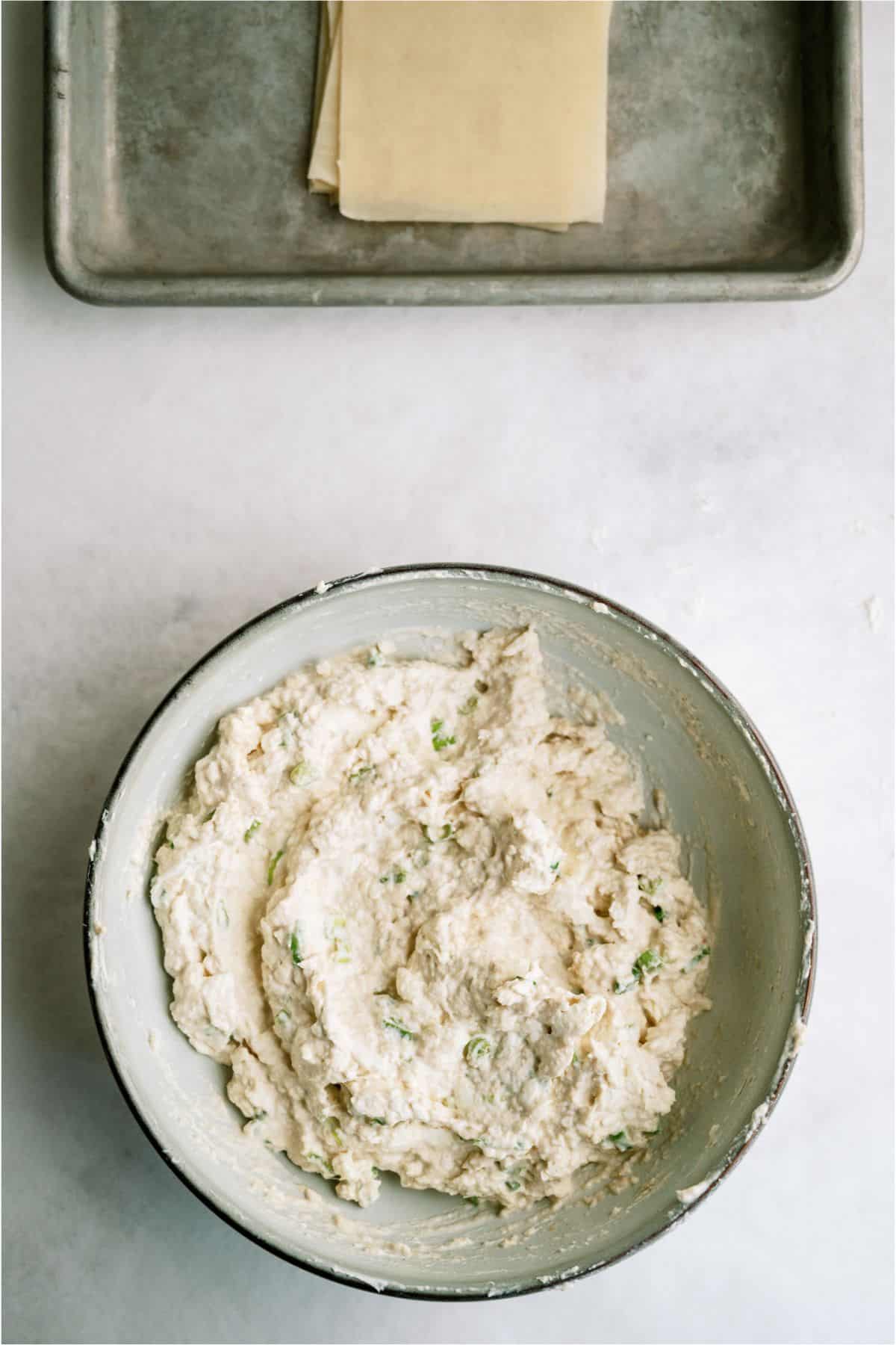 Bowl of cheese mixture with herbs on a countertop; pastry sheets on a tray in the background.