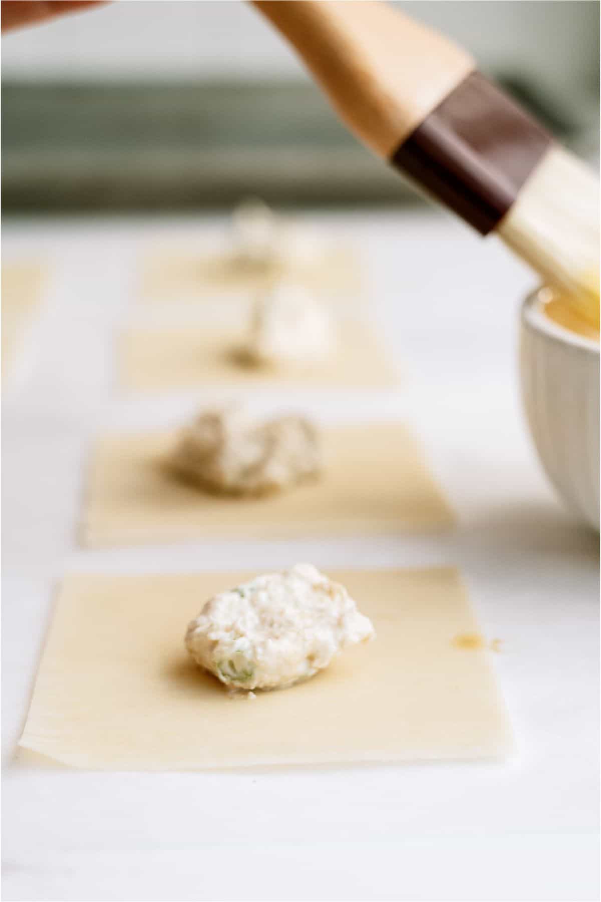 A pastry brush applies liquid to a wrapper, with dollops of filling placed on three wrappers lined up on a surface. A white bowl is partially visible on the right.
