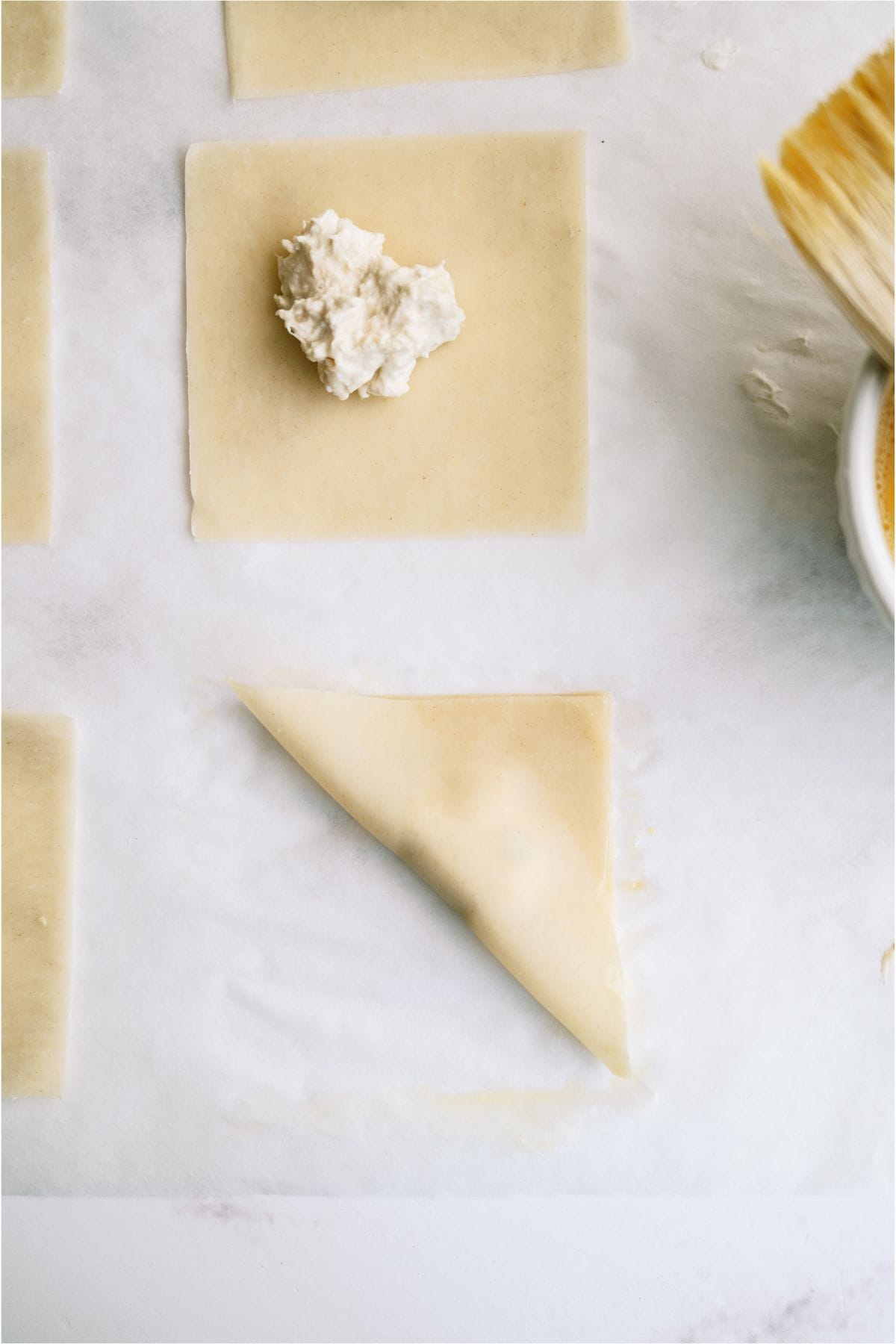 A sheet of dough with a filling placed in the center, another piece of dough folded into a triangle nearby, and a brush and bowl partially visible on the side.