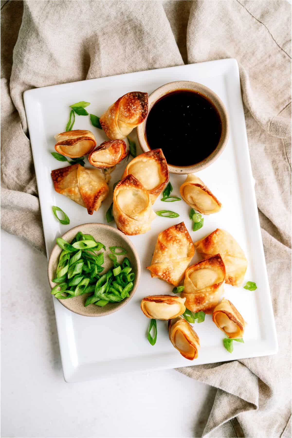 A white rectangular plate with crispy wontons, a small bowl of soy sauce, and sliced green onions on a beige cloth background.