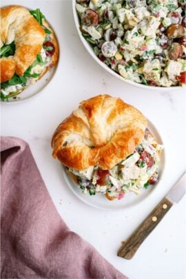 A croissant sandwich filled with chicken salad sits on a plate next to a bowl of chicken salad, a second sandwich, a cloth napkin, and a wooden-handled knife.