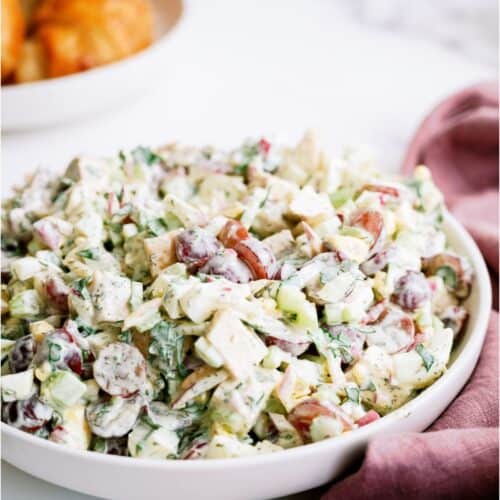 A bowl of chicken salad with grapes, celery, herbs, and a creamy dressing sits on a white surface next to a mauve napkin; croissants are blurred in the background.