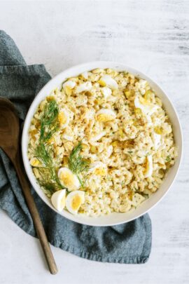 A bowl of macaroni salad with sliced boiled eggs and dill, placed on a gray cloth, with a wooden spoon beside it.