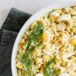 Close-up of a bowl containing egg salad garnished with fresh dill. The salad sits on a dark gray cloth, showcasing chunks of boiled egg mixed with creamy dressing.