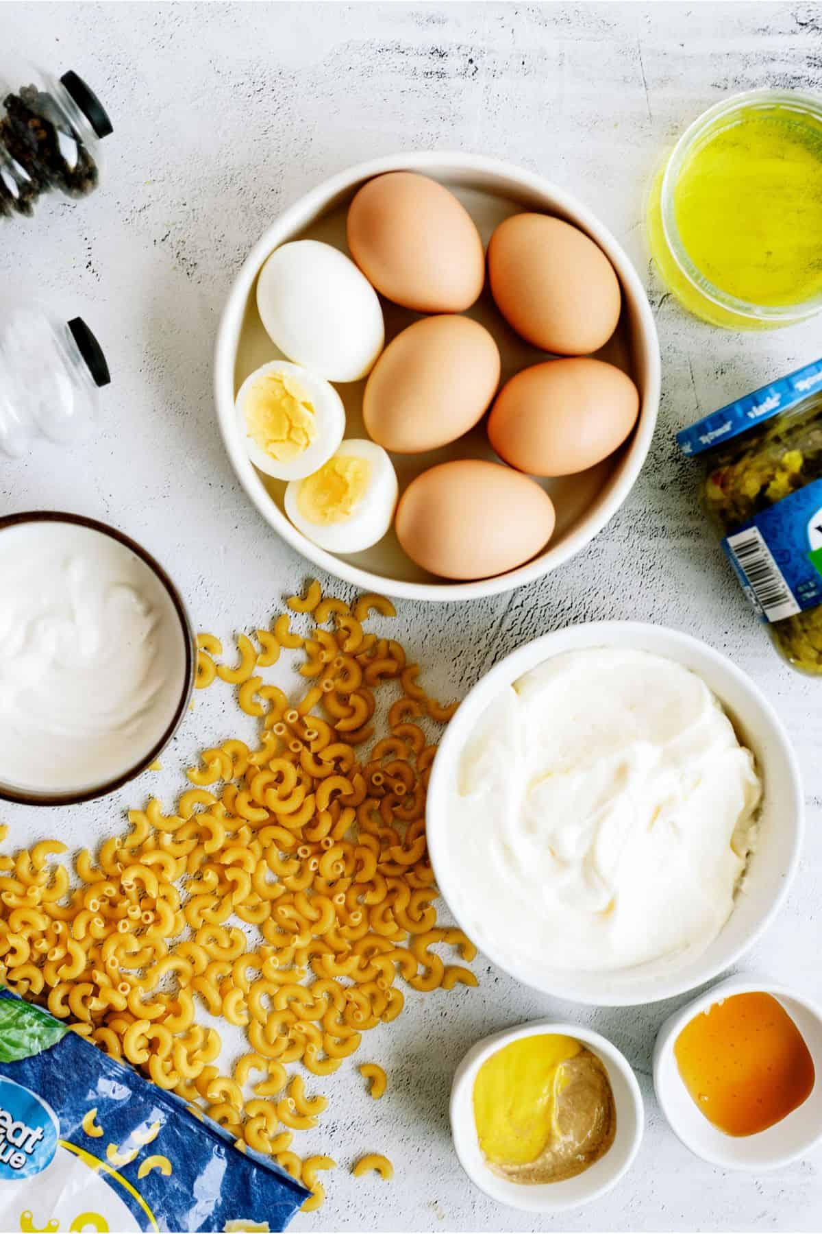 Bowls of eggs, cooked pasta, and creamy ingredients on a white surface, with mustard, pickles, and pasta surrounding them.
