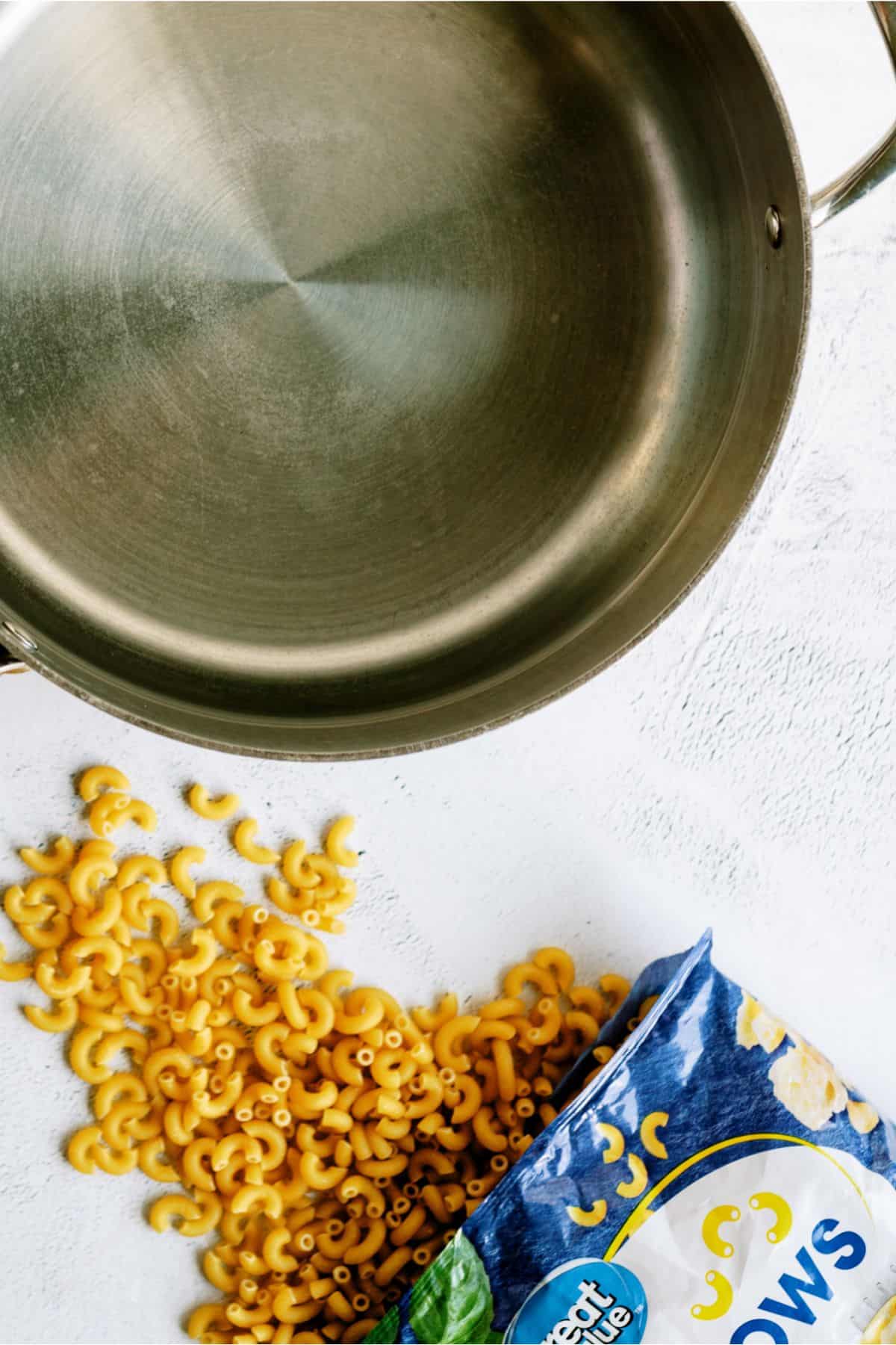 A stainless steel pot sits on a white surface next to a partially opened bag of uncooked elbow macaroni, with some pasta spilled out onto the surface.