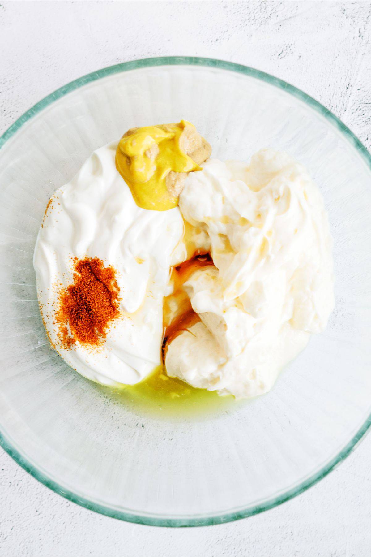 Glass bowl with yogurt, mustard, paprika, and vinegar. Ingredients are partially mixed, creating a creamy texture with distinct yellow and red seasoning atop.