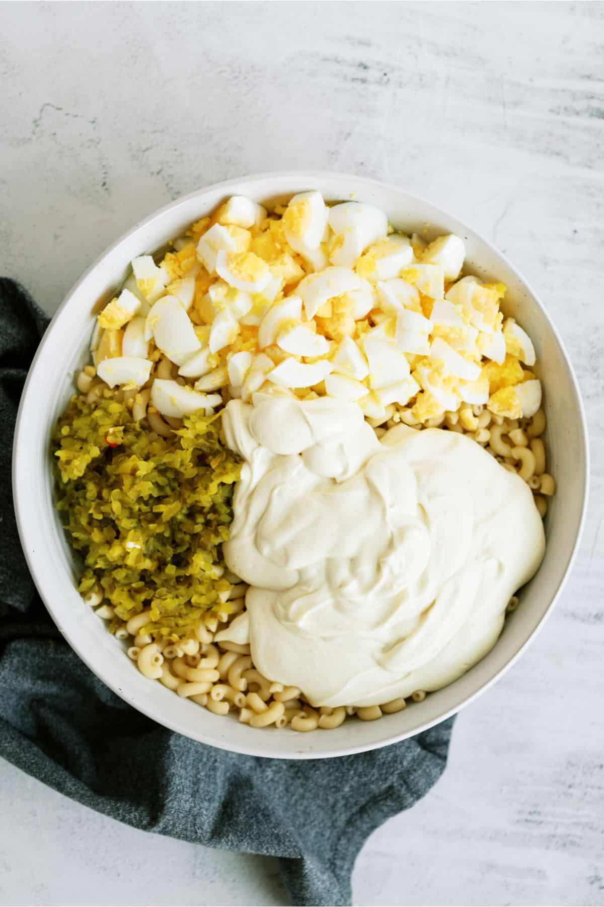 A bowl of macaroni salad ingredients: chopped boiled eggs, relish, and mayonnaise, on top of cooked macaroni, placed on a gray cloth.