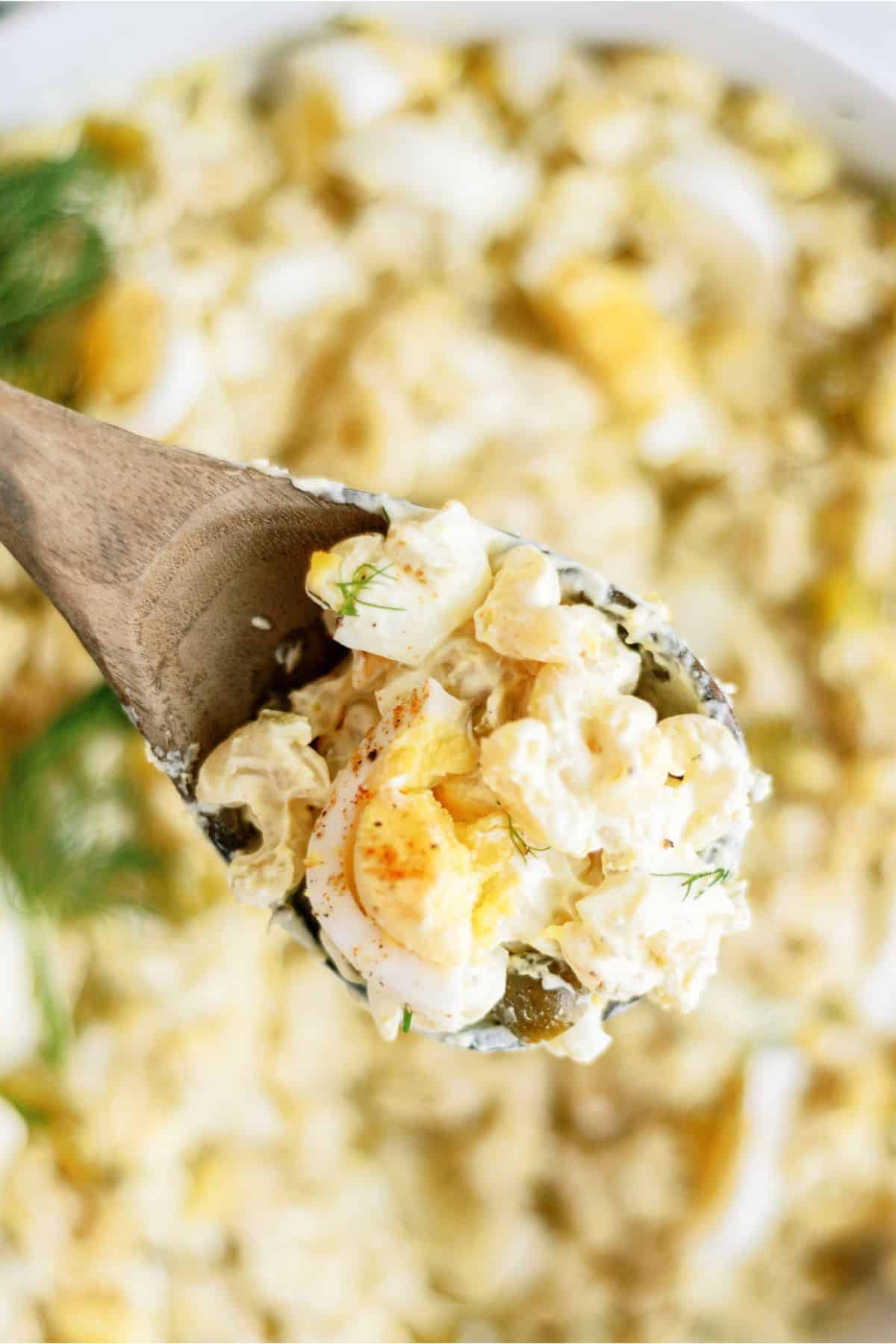 A wooden spoon holds a portion of creamy potato salad with chunks of boiled eggs, garnished with fresh herbs. The background shows more of the salad.