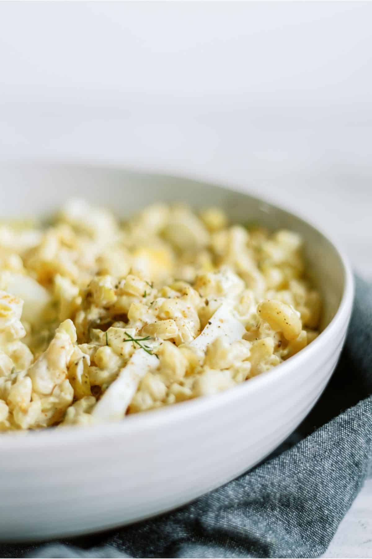 A close-up of creamy pasta in a white bowl, seasoned with herbs and pepper. The bowl rests on a gray cloth.