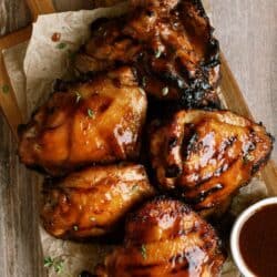 Grilled chicken thighs with a glossy barbecue glaze arranged on parchment paper atop a wooden board, accompanied by a small bowl of sauce.