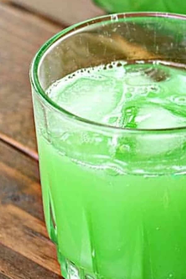 A close-up of a glass containing a green-colored beverage with ice cubes, placed on a wooden surface.