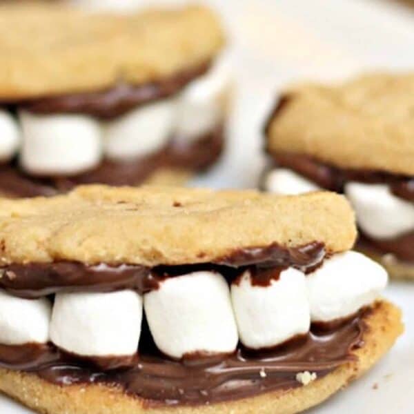 Three cookie sandwiches filled with marshmallows and chocolate, resembling smiling mouths with teeth, are placed on a white plate.