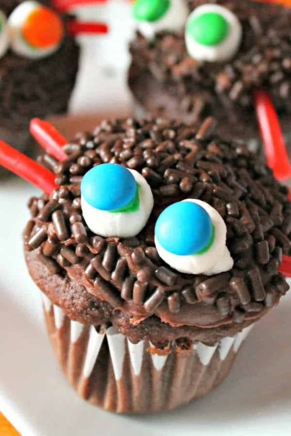 A chocolate cupcake decorated with chocolate sprinkles, two small marshmallows topped with blue and green candies resembling eyes, and red candy strips.