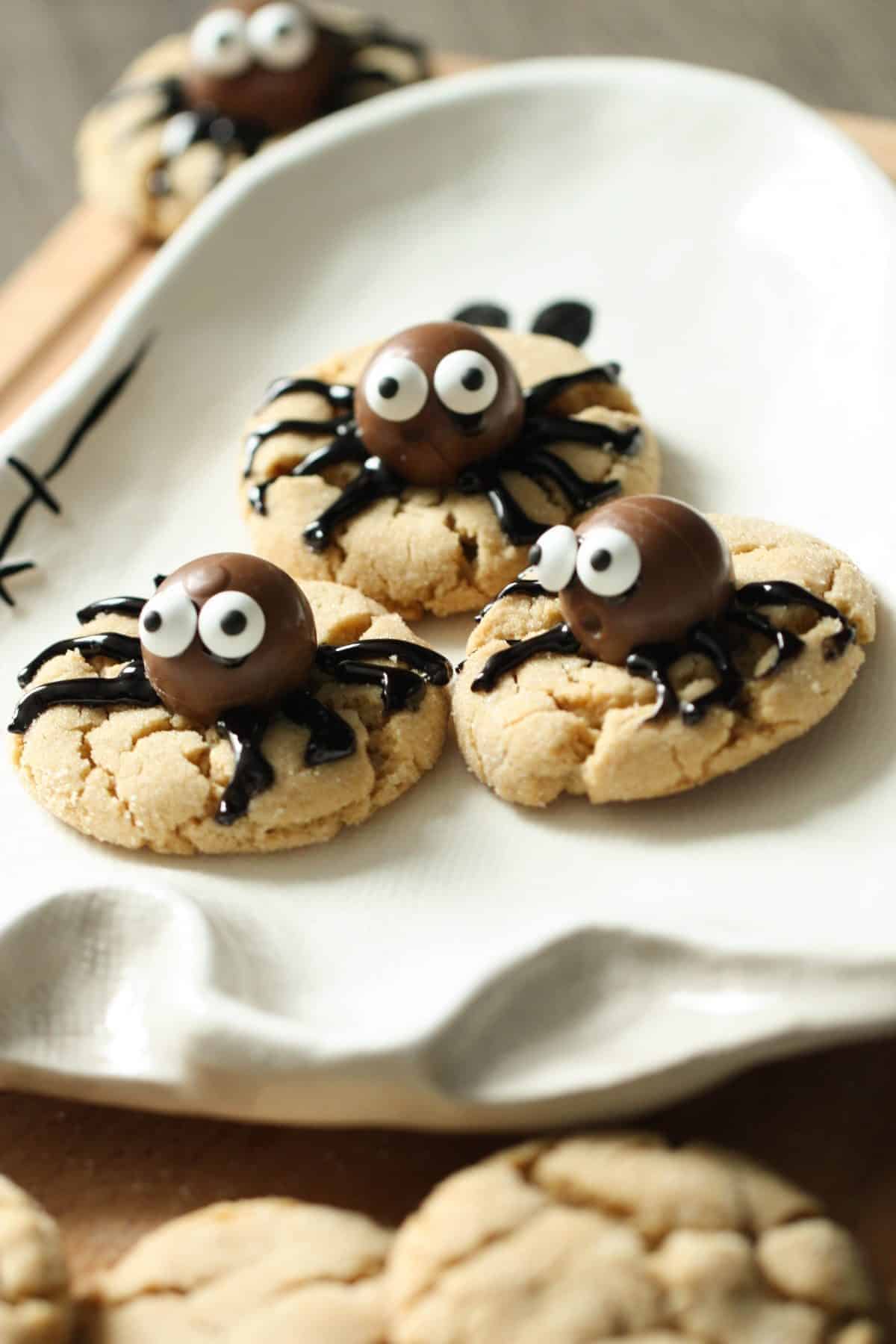 Three spider-themed cookies with chocolate candy bodies and icing legs are displayed on a white plate. Additional cookies are in the background on a wooden surface.