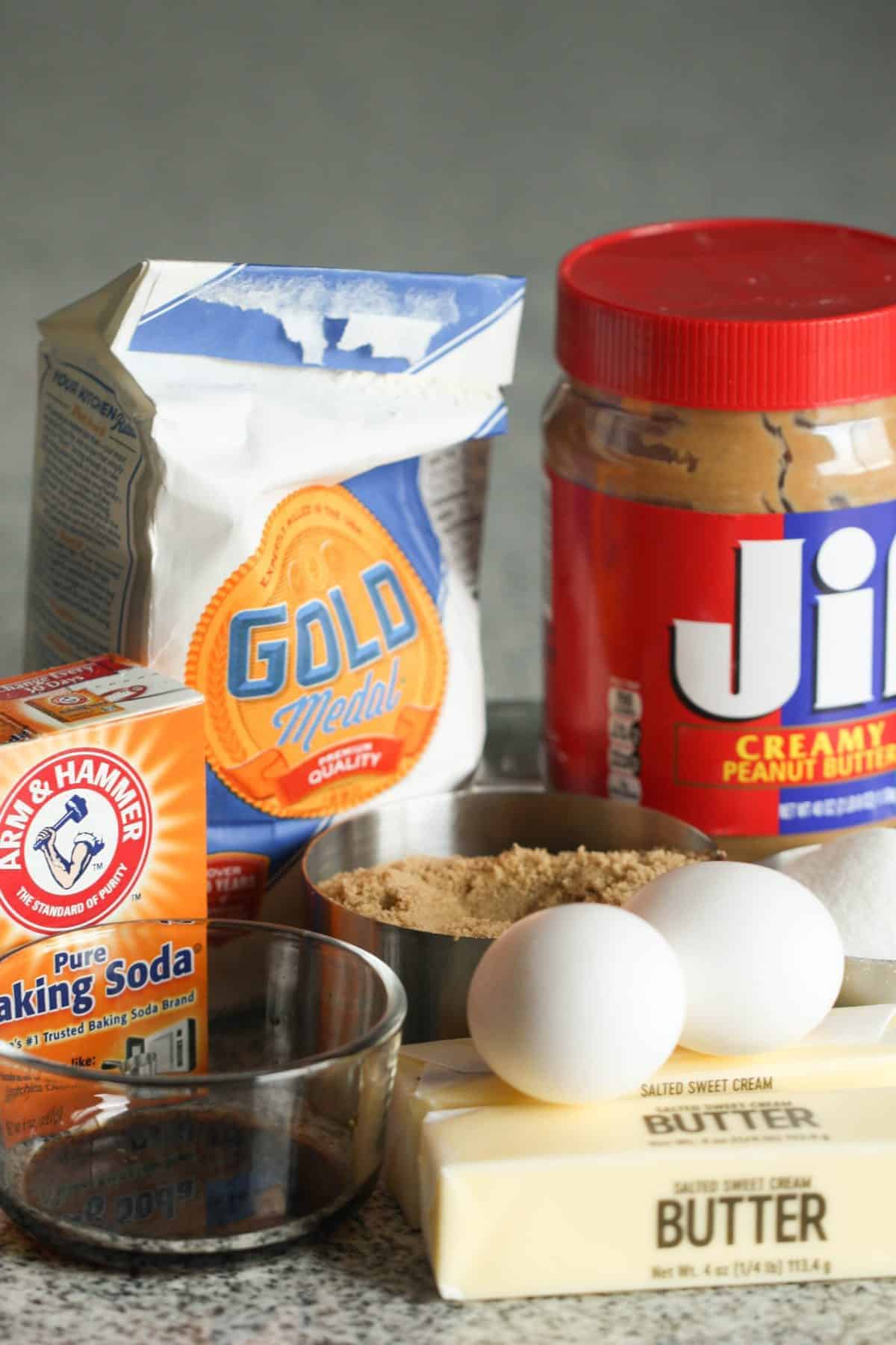 Baking ingredients including a bag of Gold Medal flour, a jar of Jif peanut butter, a box of Arm & Hammer baking soda, a stick of butter, two eggs, a cup of brown sugar, and a bowl of granulated sugar.