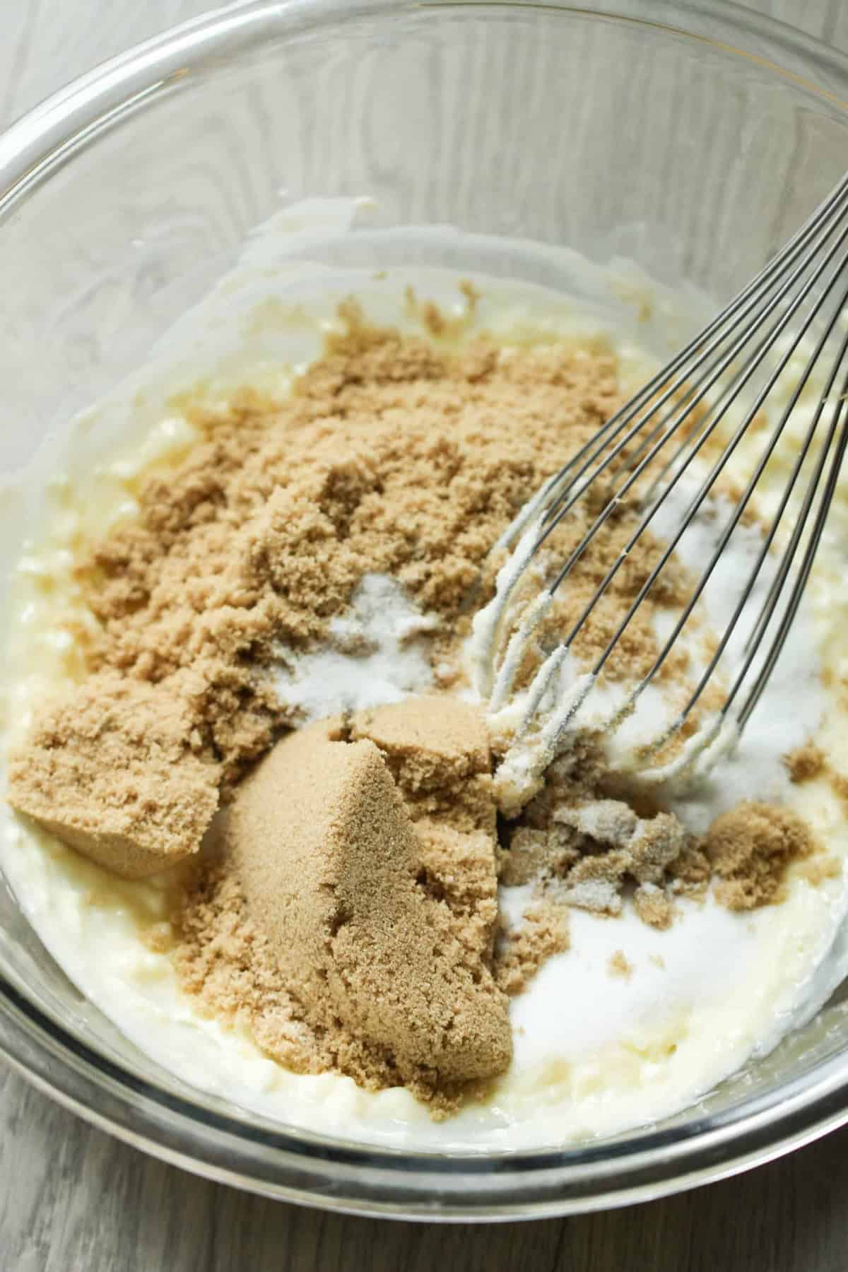 A glass bowl containing a whisk, brown sugar, and a creamy mixture on a light wooden surface.