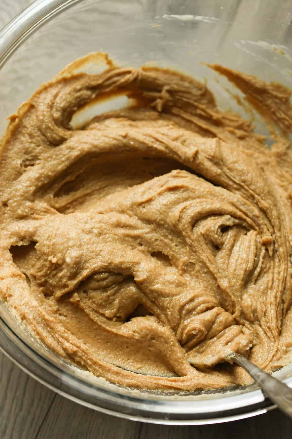 A glass bowl filled with smooth, creamy peanut butter cookie dough, with a spoon partially visible mixing the dough.