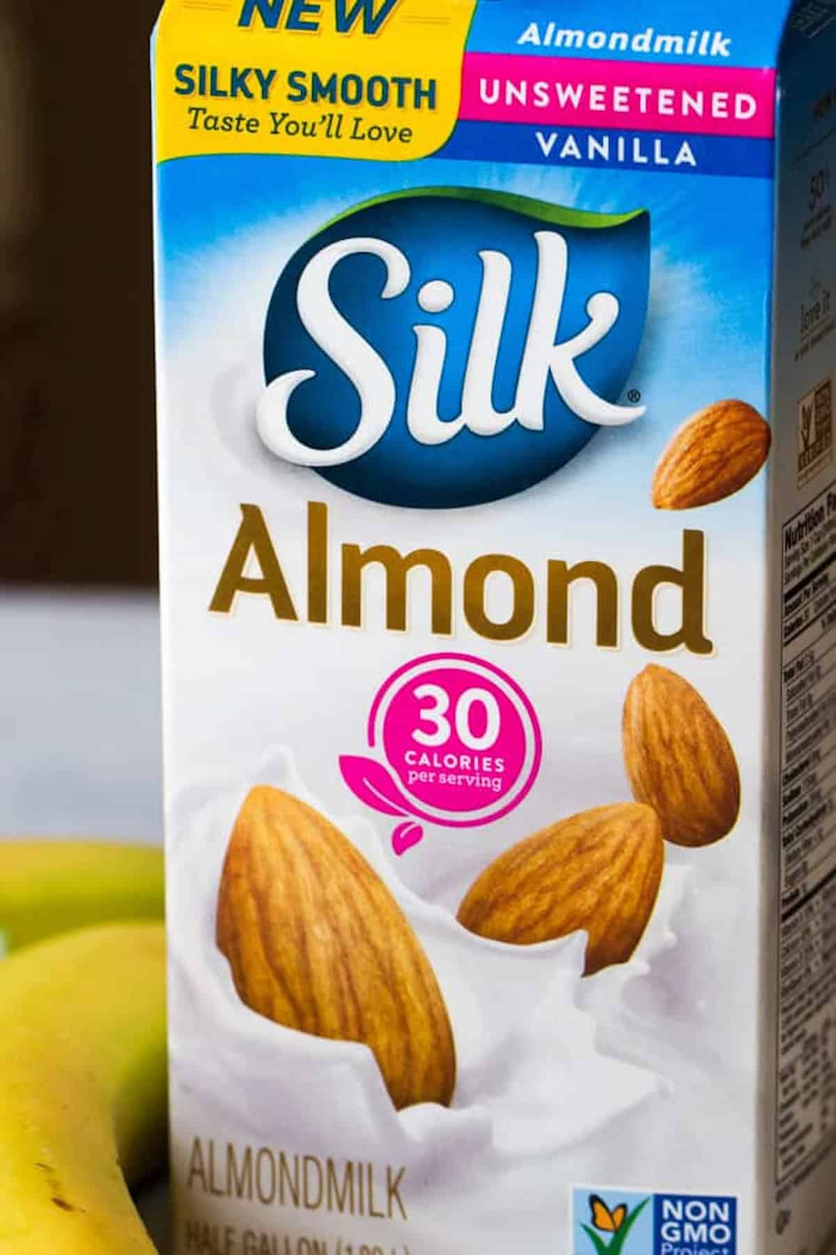 A blender cup filled with spinach, pineapple, and banana is placed next to a banana and a carton of almond milk.